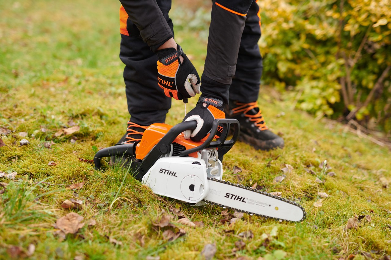 A STIHL MS 180 C-BE chainsaw on the ground, with a person wearing protective gloves holding the handle. 