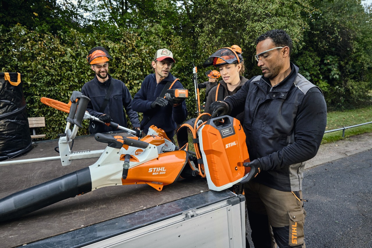 Three men and one woman wearing STIHL protective equipment are standing on a vehicle flatbed with STIHL power tools, such as the STIHL BGA 200 cordless leaf blower and the STIHL AR 2000 L backpack battery 