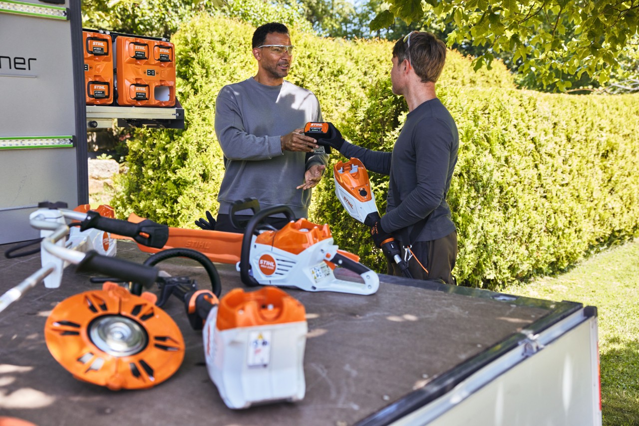 A man hands a STIHL battery to his colleague, both of whom are standing behind a vehicle flatbed, on which is, among other things, a bottTainer and a STIHL cordless chainsaw, as well as other power tools