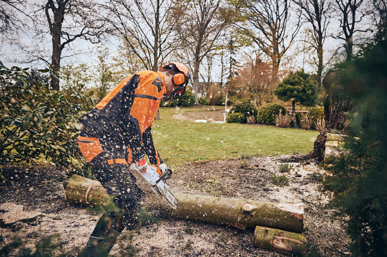 A man wearing protective equipment sawing a tree trunk in a green area with a STIHL cordless chainsaw
